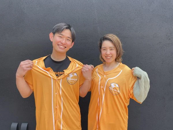 オレンジ色のオリジナルベースボールシャツを着用する男女のイメージ。スポーツチームやイベントスタッフ、応援ウェアに活用できるユニフォーム。