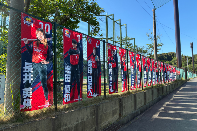 NECレッドファルコンズの選手写真をデザインしたバナーを屋外フェンス沿いに連続設置したスポーツ会場装飾事例。